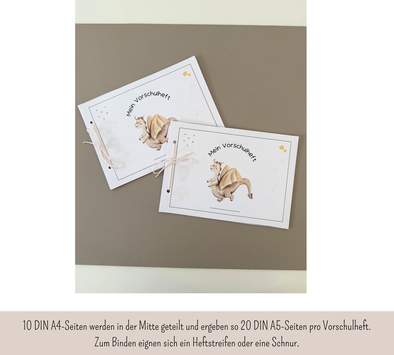 May include: Two white notebooks with the text "Mein Vorschuheft" and a cartoon dragon illustration. The notebooks are bound with a pink ribbon. The background is a solid, neutral color.