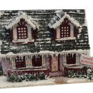 Peut inclure: Une maison miniature recouverte de neige avec un toit rouge et des bordures blanches. La maison a deux fen&ecirc;tres au dernier &eacute;tage et trois fen&ecirc;tres au rez-de-chauss&eacute;e. La maison a une pancarte qui dit "L'atelier du P&egrave;re No&euml;l".