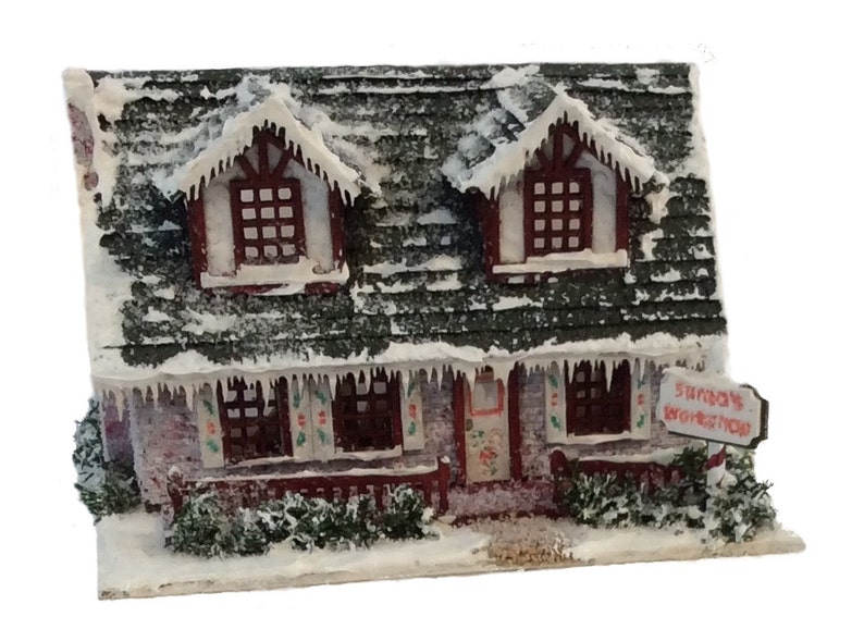 Peut inclure: Une maison miniature recouverte de neige avec un toit rouge et des bordures blanches. La maison a deux fen&ecirc;tres au dernier &eacute;tage et trois fen&ecirc;tres au rez-de-chauss&eacute;e. La maison a une pancarte qui dit "L'atelier du P&egrave;re No&euml;l".