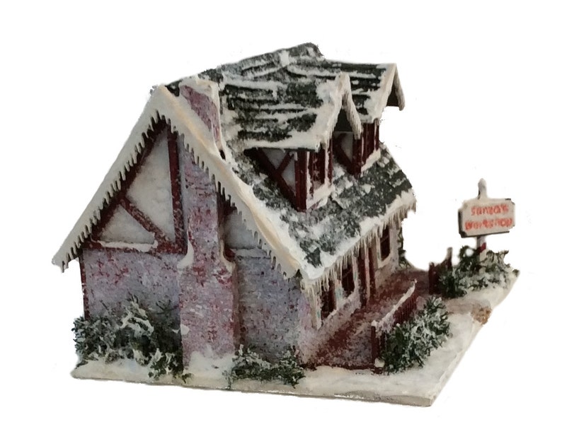 Peut inclure: Une miniature de chalet enneig&eacute; avec une chemin&eacute;e en brique rouge et une pancarte indiquant "L'atelier du P&egrave;re No&euml;l". Le chalet a un toit blanc avec des gla&ccedil;ons et une base enneig&eacute;e.