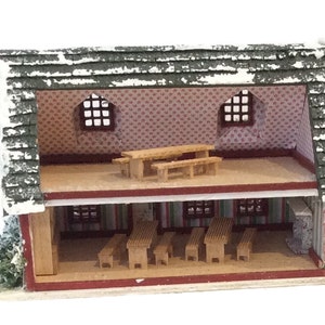 Peut inclure: Une maison de poup&eacute;e miniature en bois avec un ext&eacute;rieur rouge et blanc et un toit vert. L'int&eacute;rieur est visible, montrant une pi&egrave;ce avec des meubles en bois et un papier peint &agrave; motifs.