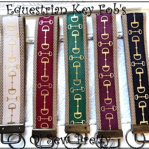 May include: Equestrian key fobs in various colors, including beige, burgundy, green, purple, and navy. Each fob features a gold-colored horse bit design on a colored ribbon, attached to a key ring. The text "Equestrian Key Fob's" is at the top.