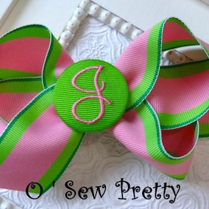 May include: A pink and green striped bow with a green button featuring a pink embroidered letter "G" in the center. The bow is on a white surface with the text "O' Sew Pretty" below it.