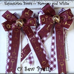 May include: Two maroon and white equestrian bows. The bows feature a gold horse bit design on maroon ribbon. The bows are accented with white and maroon gingham and white ribbon. The bows are framed in a white decorative frame.