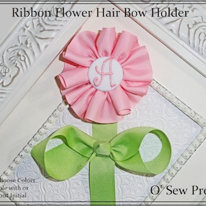 May include: A pink ribbon flower hair bow holder with a green ribbon bow and a white button with the letter "A" embroidered on it. The holder is displayed against a white background with a decorative border.