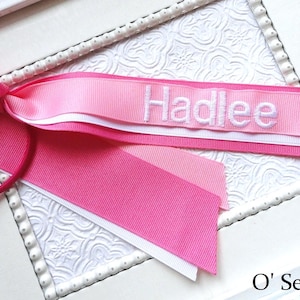May include: A pink hair bow with the name "Hadlee" embroidered in white. The bow features a layered design with shades of pink and white, and includes a matching pink hair tie.