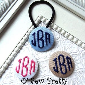 May include: Three personalized hair ties with the initials "JBA" embroidered on fabric-covered buttons. One is light blue with navy embroidery, one white with pink, and one tan with navy. The text "O' Sew Pretty" is at the bottom.