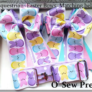 May include: Easter-themed equestrian bows and matching belt. The bows and belt are white with a repeating pattern of colorful Easter bunny designs in pink, purple, yellow, and blue. The belt has a silver buckle.