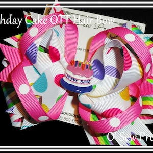 Puede incluir: Un lazo para el pelo rosa y blanco con un dije de pastel de cumplea&ntilde;os. El lazo est&aacute; hecho de cinta con lunares blancos y rayas de colores. El dije es un pastel peque&ntilde;o con velas y las palabras "Happy B.Day".