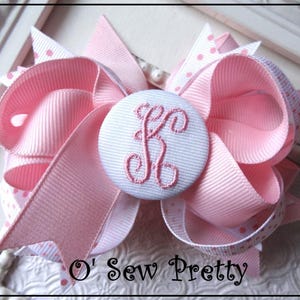 May include: A pink and white polka dot hair bow with a white button featuring an embroidered letter "F". The bow is made of grosgrain ribbon and has a layered design. The text "O' Sew Pretty" is visible at the bottom of the image.