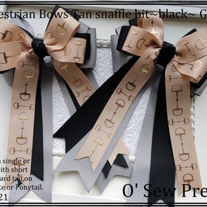 May include: Two equestrian bows with tan, black, and gray ribbon. The ribbon has a gold snaffle bit design. The bows are sold as a single or a pair, with short or standard tail, on a barrette or ponytail. "O' Sew Pretty" is written on the bottom right corner of the image. 2/22/21 is written on the bottom left corner of the image.