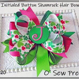 May include: Green, white, and pink shamrock patterned hair bow with a green button with a white letter J in the center. The bow is on a white background with a decorative border. The text "O' Sew Pretty" is at the bottom of the image.