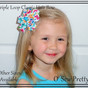 May include: A young girl with blonde hair is wearing a blue shirt and a colorful butterfly hair bow. The bow has a white background with multi-colored butterflies. The text "Triple Loop Classic Hair Bow" and "O' Sew Pretty" are visible in the image.