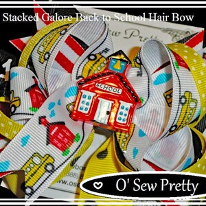May include: Triple stacked hair bow featuring a schoolhouse charm. The bow is made from white, yellow, red and black ribbon with a school bus print. The charm is red with white windows and a yellow door. The text "SCHOOL" is printed on the charm. The bow is tied with a yellow ribbon and has a heart-shaped tag with the text "O' Sew Pretty".