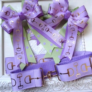 May include: Three purple and green ribbon bows with gold equestrian stirrup designs. The bows are attached to a purple strap with a gold buckle.