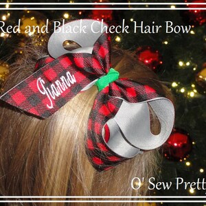 May include: Red and black plaid hair bow with the name "Gianna" embroidered in white. The bow is tied with a green ribbon and is shown on a child's hair.