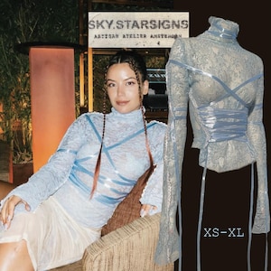 May include: A light blue lace top with long sleeves and a high neck, featuring blue ribbon accents. The top is displayed on a mannequin and worn by a person. The text "XS-XL" is visible. The background includes a sign that reads "SKY.STARSIGNS ARTISAN ATELIER AMSTERDAM".
