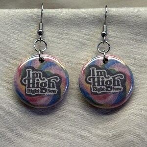 May include: A pair of round earrings with a colorful, swirled background. The earrings feature the text "I'm High Right Now" in a stylized font. The earrings have silver-colored hooks.