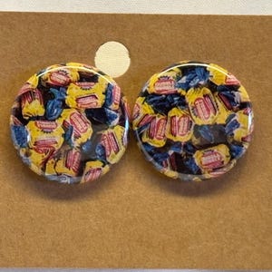 May include: A pair of round stud earrings. Each earring features a clear resin dome filled with miniature bubble gum wrappers in yellow, blue, and red. The earrings are displayed on a brown card.