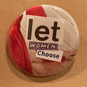 May include: A round button with a red, white, and wood-grain background. The button has a white paper sticker with the text "let women choose" in black letters.