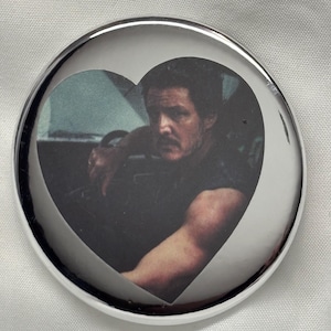May include: A round, silver-colored button with a heart-shaped cutout revealing a portrait of a man. The man is dark-haired and appears to be driving a vehicle. The button has a shiny, reflective surface.