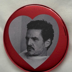 May include: A red button with a heart-shaped cutout featuring a black and white image of a man with dark hair and a mustache. The button has a glossy finish and a metallic appearance.