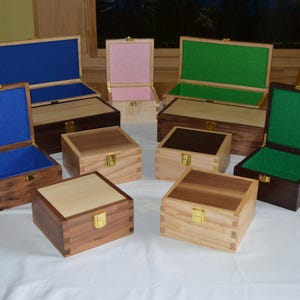 Peut inclure: Plusieurs boîtes en bois avec des couvercles à charnières, certaines ouvertes révélant des doublures en feutre bleu, rose et vert. Les boîtes varient en taille et en teinte de bois, avec des fermoirs dorés. Elles sont sur une nappe blanche.