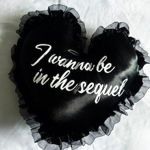 Può includere: Un cuscino nero a forma di cuore con la scritta "I wanna be in the sequel" in bianco. Il cuscino ha un bordo a balze sui bordi. Il cuscino è su una superficie bianca.
