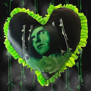 May include: Heart-shaped black pillow with a green-tinted face print and a neon green ruffled border. The pillow features a circular logo and is set against a dark background with green thorn accents.