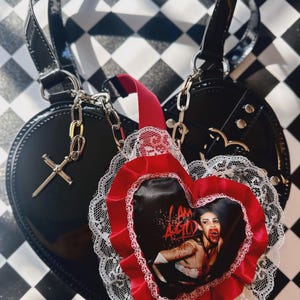 May include: Black heart-shaped handbag with silver chain straps and a cross charm. A red and white heart-shaped accessory with a vampire image and the text "I AM A GOTH" is attached to the bag. The background is a black and white chequered pattern.
