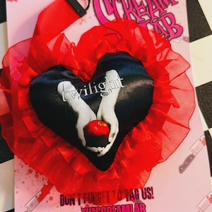 May include: A red lanyard with a black clasp and a heart-shaped charm. The charm features a black background with the word "twilight" and an image of hands holding a red apple. The charm is surrounded by red ruffled fabric.