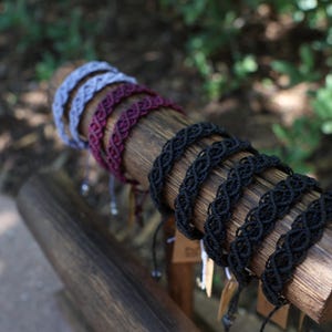 May include: A collection of woven bracelets in various colors, including black, gray, and burgundy, displayed on a wooden log. The bracelets have an intricate braided design and adjustable closures. The background is blurred with green foliage.