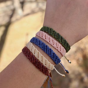 May include: A collection of macrame bracelets in various colors, including burgundy, cream, blue, pink, and green. Each bracelet features a woven design with a small opening. The bracelets are displayed on a wrist.