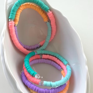 Könnte beinhalten: Ein Set aus vier bunten Armbändern aus übereinander gestapelten, runden Perlen. Die Armbänder sind in den Farben Rosa, Orange, Lila und Türkis gehalten. Die Perlen sind mit kleinen, klaren Kristallen durchsetzt.