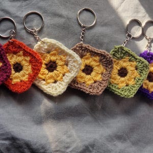 Può includere: Sei portachiavi a forma di girasole all'uncinetto con diversi sfondi colorati. I portachiavi sono viola, arancio, bianco, marrone, verde e viola.