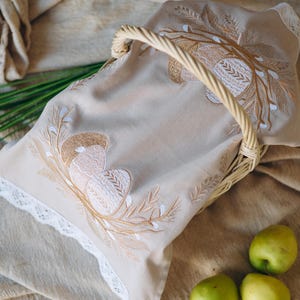 May include: A beige linen basket with a woven handle, holding a matching embroidered cloth. The cloth features a detailed design of stylized eggs and branches in shades of brown and white. Three green apples rest nearby.