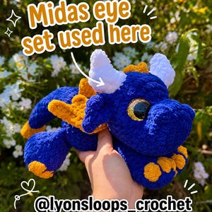 Op de afbeelding: Een blauwe gehaakte drakenknuffel met witte hoorns, gele vleugels en gele accenten op de voeten. Het speelgoed heeft grote, glinsterende gouden ogen en bevat de tekst Midas eye set used here en @lyonsloops_crochet op de afbeelding.