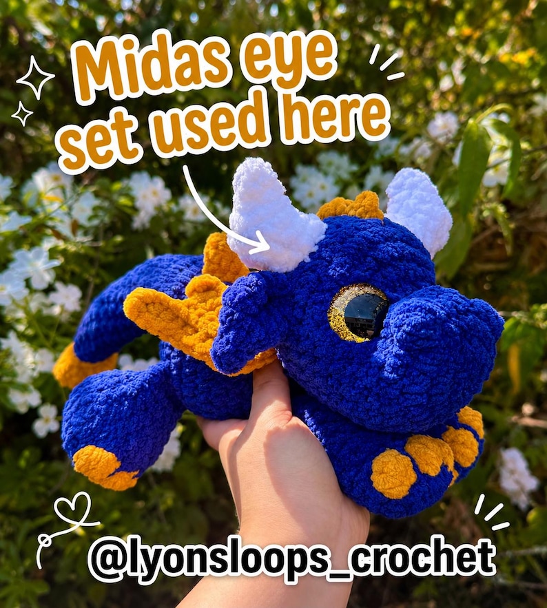 Op de afbeelding: Een blauwe gehaakte drakenknuffel met witte hoorns, gele vleugels en gele accenten op de voeten. Het speelgoed heeft grote, glinsterende gouden ogen en bevat de tekst Midas eye set used here en @lyonsloops_crochet op de afbeelding.