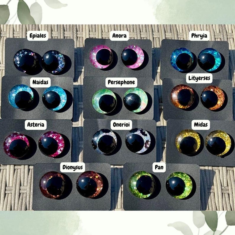 Op de afbeelding: Een collectie van tien paar ronde, bolvormige knutselogen gemonteerd op zwarte kaarten, elk gelabeld met namen zoals Epiales, Anora, Phryia, Naidas, Persephone, Lityerses, Asteria, Onerioi, Midas, Dionysus en Pan, in diverse glitterende kleuren.