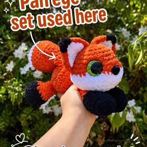 Op de afbeelding: Een gehaakt vossenknuffeltje van oranje, wit en zwart garen, met grote groene ogen en een pluizige staart. De afbeelding bevat de tekst 'Pan eye set used here' met een pijl naar de vos en '@lyonsloops_crochet' onderaan.