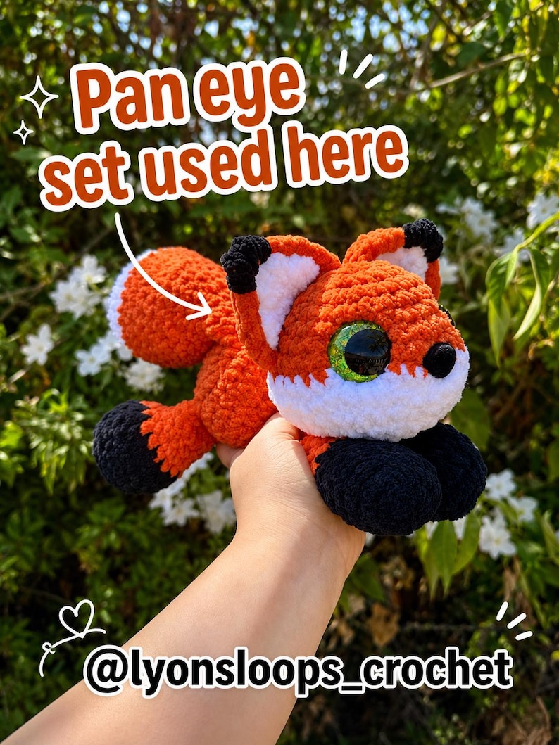 Op de afbeelding: Een gehaakt vossenknuffeltje van oranje, wit en zwart garen, met grote groene ogen en een pluizige staart. De afbeelding bevat de tekst 'Pan eye set used here' met een pijl naar de vos en '@lyonsloops_crochet' onderaan.