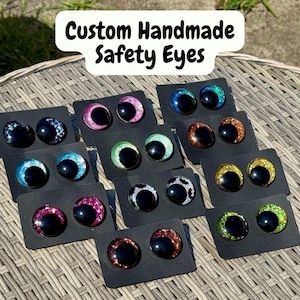 Op de afbeelding: Een collectie handgemaakte veiligheidsoogjes voor knuffels, elk met een grote zwarte pupil omringd door een kleurrijke, glitterende iris. De paren zijn bevestigd op individuele zwarte kaartjes en tonen diverse kleuren zoals roze, blauw, groen, oranje, geel en zilver.