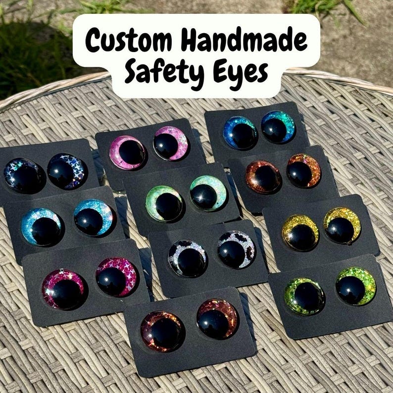 Op de afbeelding: Een collectie handgemaakte veiligheidsoogjes voor knuffels, elk met een grote zwarte pupil omringd door een kleurrijke, glitterende iris. De paren zijn bevestigd op individuele zwarte kaartjes en tonen diverse kleuren zoals roze, blauw, groen, oranje, geel en zilver.