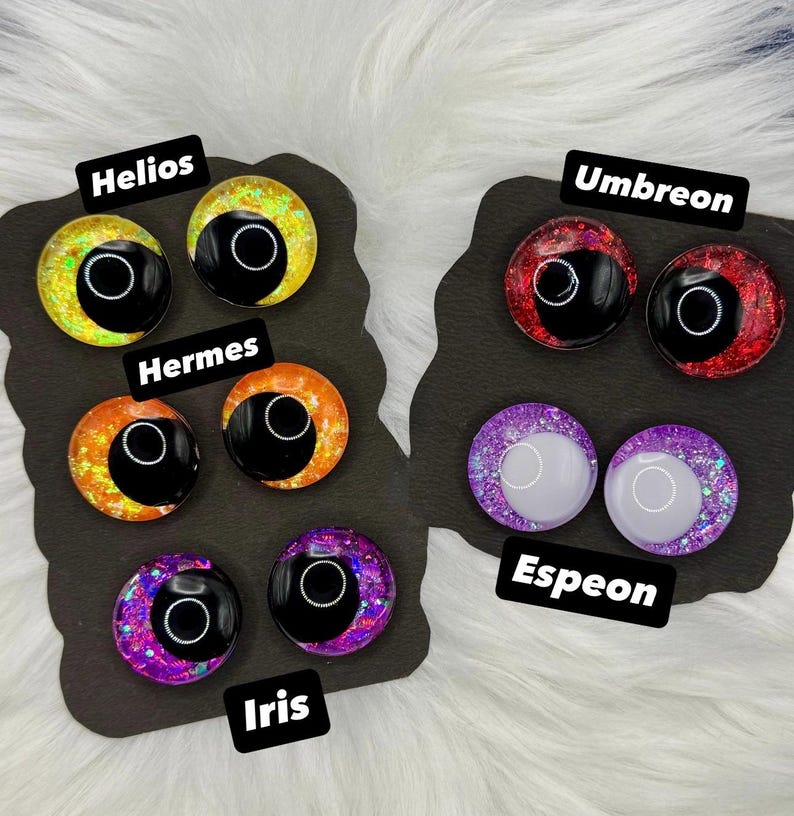 Op de afbeelding: Zes paar ronde hars cabochons met glitterende, veelkleurige irissen en grote zwarte pupillen. De sets zijn gelabeld als Helios, Hermes, Iris, Umbreon en Espeon, weergegeven op twee zwarte kaarten met een glanzende, reflecterende afwerking.