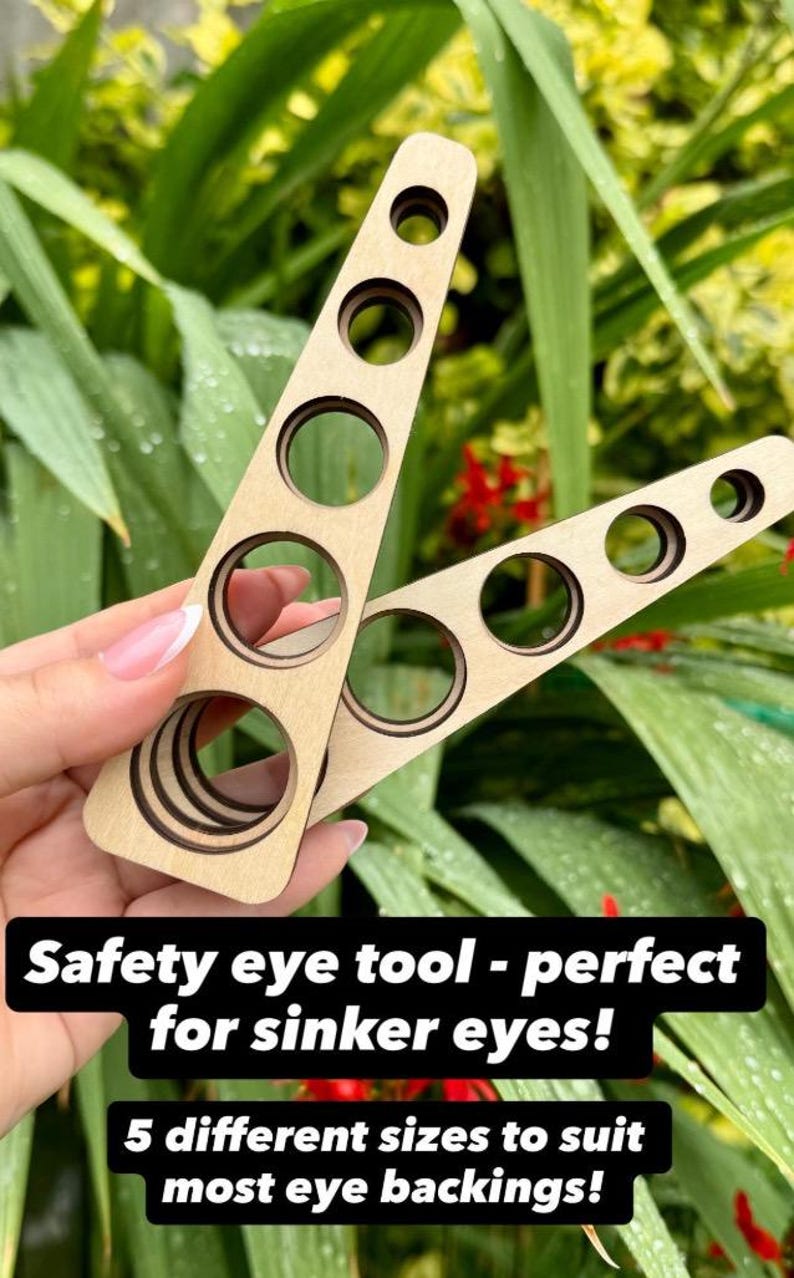 Op de afbeelding: Een veiligheidsoog-gereedschap van lichtgekleurd hout, bestaande uit twee driehoekige delen met vijf ronde uitsparingen van verschillende diameters. De tekst op de afbeelding luidt: Safety eye tool - perfect for sinker eyes! 5 different sizes to suit most eye backings!