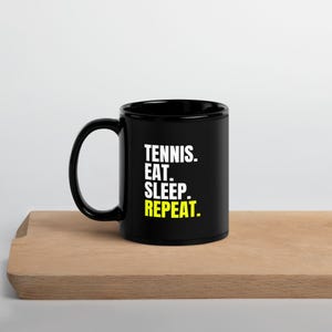 May include: Black ceramic coffee mug with white text that reads "Tennis. Eat. Sleep. Repeat." The text "Repeat" is in yellow.