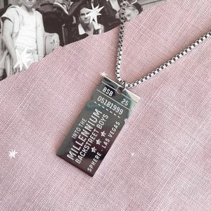 Puede incluir: Un collar con colgante rectangular plateado. El colgante tiene el texto "Into the Millennium Backstreet Boys Sphere Las Vegas" y los números "BSB 05181999 25". El collar está sobre una superficie rosa.