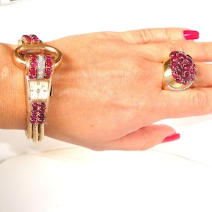 Puede incluir: Un juego de reloj de pulsera y anillo de oro con rubíes y diamantes. El reloj tiene una esfera rectangular y una correa adornada con rubíes. El anillo presenta un grupo de rubíes engastados en una banda de oro con detalles de diamantes.