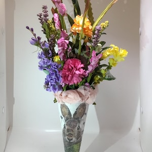 May include: A colorful bouquet of flowers in a cone-shaped vase with a bunny rabbit design. The flowers include pink, purple, yellow, and orange.