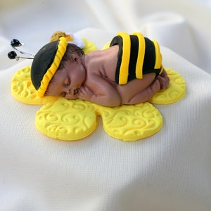 May include: A small, sleeping baby figurine made of fondant, dressed in a yellow and black bee costume, lying on a yellow flower-shaped fondant base.